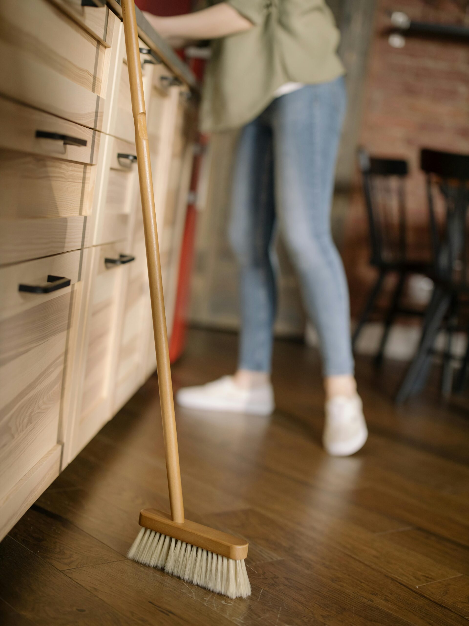 A woman in casual attire sweeps the kitchen floor, maintaining cleanliness in a cozy home.